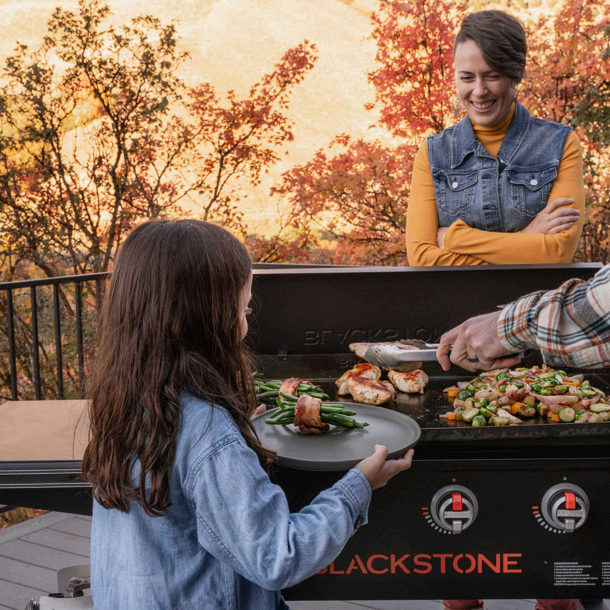 Perhe kokkaamassa ulkona Blackstone 36" ParilaGrillillä kovalla kannella syksyllä, nauttien ateriasta värikkään syyslehvästön ympäröimänä.